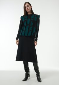 Grüner und schwarzer gemusterter Pullover mit vertikalen Designelementen, kombiniert mit einem schwarzen Midikleid und knielangen schwarzen glänzenden Stiefeln.