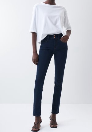 Person iført hvid løs t-shirt, mørkeblå slim-fit jeans med to knapper og sorte sandaler med hæl og stropper mod en ensfarvet baggrund.
