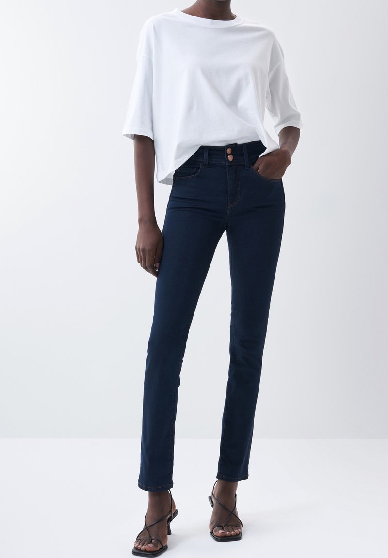 Personne portant un t-shirt blanc ample, un jean slim bleu foncé à deux boutons, et des sandales à talons noires à brides sur un fond uni.