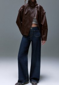 Personne portant une veste en cuir marron, un top court blanc, un jean large bleu foncé et des baskets grises sur un fond gris uni.