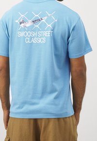 Modré bavlněné tričko s kulatým výstřihem. Na zadní straně je bílé logo "Swoosh" a text "SWOOSH STREET CLASSICS" na mřížkovaném vzoru.