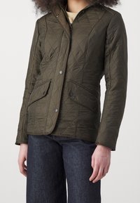 Quilted olive green jacket with snap buttons, two front pockets, and a high collar. Features a diamond pattern on the fabric.