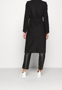Persona vestida con un abrigo negro ceñido, pantalones de cuero negros y zapatillas blancas, de pie contra un fondo claro y liso.