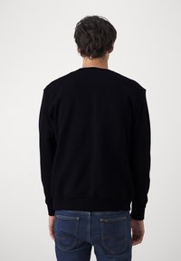 Man met donker haar, gekleed in een zwarte trui met lange mouwen en een blauwe spijkerbroek, staat met de rug naar de camera tegen een effen lichte achtergrond.