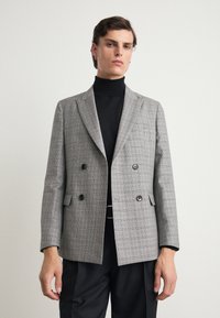 Grå rutig dubbelknäppt blazer med slagkragar, två svarta knappar och en bröstficka, bärs över en svart polotröja.