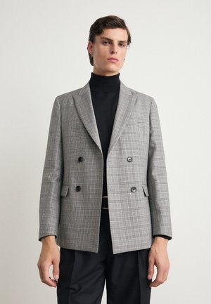 Grijze geruite dubbelgeknoopte blazer met notch revers, twee zwarte knopen en een borstzak, gedragen over een zwarte coltrui.