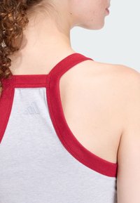 Szary sportowy top na ramiączkach z czerwonymi wykończeniami, z tylną częścią w stylu racerback oraz małym logo z trzema paskami na plecach. Miękka faktura.
