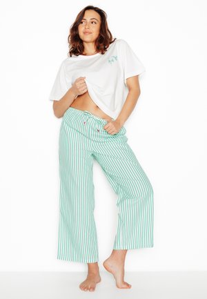 Femme debout pieds nus, soulevant un t-shirt blanc pour montrer le ventre, portant un pantalon ample à rayures vertes et blanches avec un cordon de serrage.