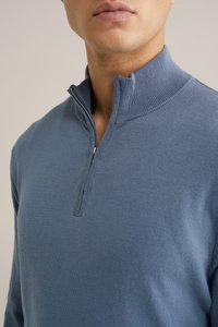 Kék kötött pulóver, cipzáras nyakkal, textúrált gallérral és sima testtel. Az anyag lágy és közepes súlyú. Minták nem láthatók.