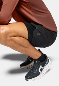 Chaussures de sport noires avec des accents blancs et un matériau texturé, associées à un short foncé. Un haut bordeaux est partiellement visible. Les chaussettes sont noires.
