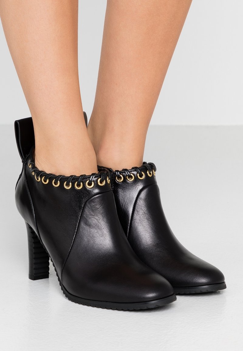 Botines de cuero negro hasta el tobillo con puntera puntiaguda, parte superior texturizada, ojales dorados y un alto tacón cuadrado. Costuras detalladas en el cuello.