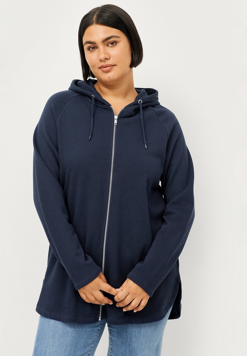 Marine blauwe zip-up hoodie met een trekkoordcapuchon, lange raglanmouwen, zijslitsen en een zachte textuur. Draag het over lichtblauwe jeans.