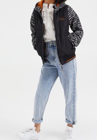 Giacca nera trapuntata con maniche a strisce zebra e foderato arancione, indossata sopra una maglietta bianca e jeans blu arrotolati, abbinata a sneakers bianche.