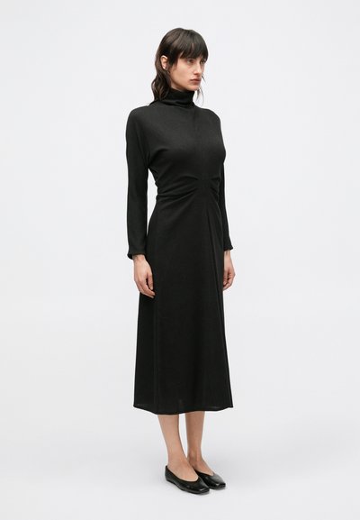 Fekete hosszú ujjú ruha magas nyakkal, testhez simuló felsőrésszel és bővülő szegéllyel. Texturált anyagból készült, derékvonalban sliccelt részletekkel.
