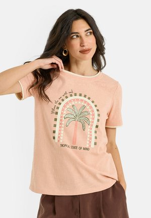 Femme aux longs cheveux foncés, portant des boucles d'oreilles créoles et un t-shirt couleur pêche avec un palmier et le texte « Tropical State of Mind ».