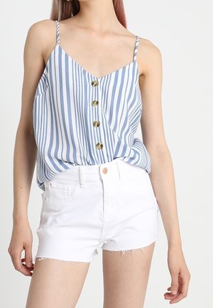 Mujer con una blusa sin mangas a rayas verticales en azul y blanco con botones y pantalones cortos de mezclilla blancos con dobladillo deshilachado.