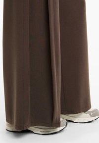 Braune, weit geschnittene Hosen aus weichem Stoff, mit glatter Textur und sauberen Säumen; kombiniert mit hellen Sneakers mit strukturierten Sohlen.