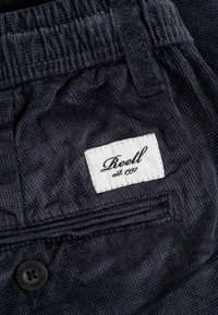 Dark blue corduroy fabric pants with a textured surface, featuring a white label with "Reell est. 1997" and a black button detail.