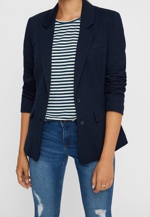 Femme portant un blazer bleu marine à manches retroussées sur une chemise rayée noire et blanche et un jean bleu déchiré.