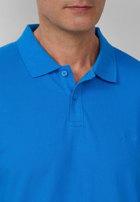Niebieska koszulka polo z teksturowanej tkaniny, z kołnierzykiem i zapięciem na dwa guziki, oraz małym logo na piersi.
