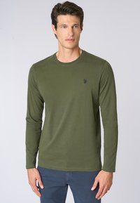 Camicia a maniche lunghe verde oliva in stoffa morbida, con scollatura rotonda e un sottile dettaglio del logo sul petto. Texture liscia.