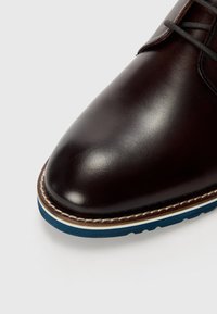 Dunkelbraune Lederschuhe mit runder Zehenpartie, einer kontrastierenden blauen Sohle, weißen Nähten und glatter Textur.