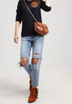 Mujer con suéter negro de punto tipo cable, jeans azules claros rotos, botas marrones con flecos hasta el tobillo y bolso cruzado de cuero marrón con borla.