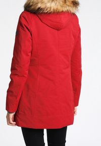 Rød vandtæt parka med en pelsforet hætte, lange ærmer og en tætsiddende pasform. Har synlige syninger og et accent-ikon på ærmet.