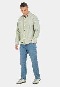 Camisa de botones verde claro con una textura suave, mangas largas y botones oscuros de contraste, combinada con jeans de mezclilla azul claro y zapatillas grises.