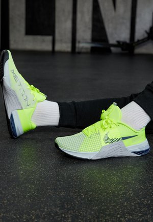 Person iført neon gule sportssko og hvide sokker, der sidder på fitnessgulvet med udstrakte ben.