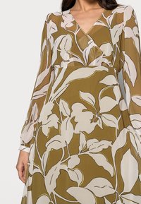 Pitkähihainen mekko oliivinvihreänä, jossa on valkoisia ja kultaisia kukkakuoseja. Mekossa on syvä V-kaula-aukko ja vyötäröä korostava muotoilu.