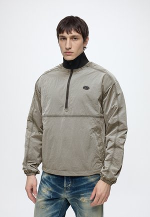 Homme portant un coupe-vent gris clair zippé avec un col noir haut et un jean bleu usé, debout devant un fond blanc uni.