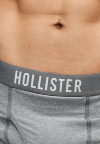 Gros plan sur le bas-ventre d'une personne avec un anneau de nombril et une ceinture de sous-vêtement Hollister grise visible.