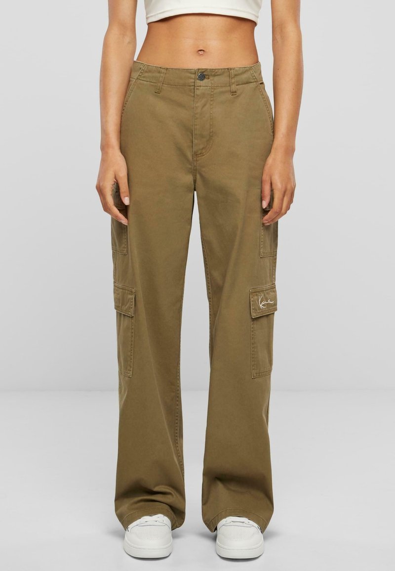 Femme portant un pantalon cargo taille haute, jambes larges, vert olive avec poches à rabat et baskets blanches, sur un fond gris uni.