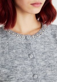 Gray knit cardigan featuring a decorative beaded collar, round buttons, and a soft texture. Detailed stitching adds visual interest.