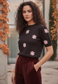 Kobieta z kręconymi włosami, ubrana w ciemną bluzkę w różowe wzory kwiatowe oraz spodnie w kolorze burgundowym o wysokim stanie, stojąca na zewnątrz wśród jesiennych liści.