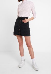 Gonna in denim nera con taglio dritto e bottoni frontali. Indossata con un top a maniche lunghe a fantasia rosa chiaro e sneakers bianche.