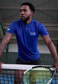 Blå piké med logo, kortermet og krage. Iført sorte shorts og holder en tennisracket med et lys grønt snorsmønster.