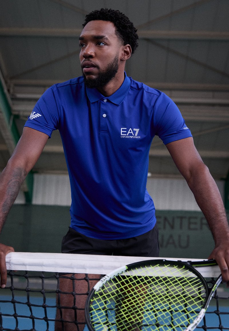 Blå piké med logo, kortermet og krage. Iført sorte shorts og holder en tennisracket med et lys grønt snorsmønster.