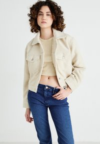 Cream-colored, textured, cropped jacket with a collar and button front, paired with a beige top and dark blue, fitted jeans.