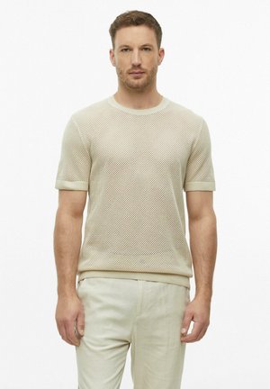 Mann trägt beige kurzärmeligen Mesh-Strickpullover und hellbeige Hose, steht vor weißem Hintergrund.