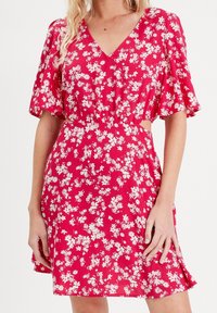 Robe florale rouge avec des manches courtes à volants, décolleté en V, taille cintrée et motifs de fleurs blanches, présentant des découpes sur les côtés.