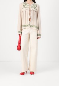 Blusa beige con bordado floral verde, con escote atado; combinada con pantalones de pierna ancha en crema y zapatos de tacón rojos. Bolso rojo sostenido en la mano.