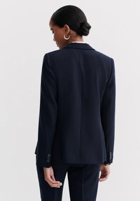 Blazer blu scuro con una vestibilità su misura, rever a punta e polsini con bottoni, caratterizzato da una texture liscia e una silhouette strutturata.
