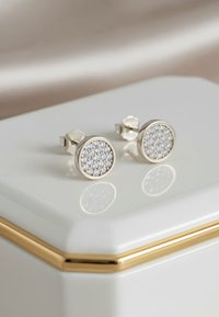 Silverfärgade studs med rund design, prydda med en klunga av små klara stenar som är inlagda i en strukturerad yta, presenterade på en vit hexagonal låda.