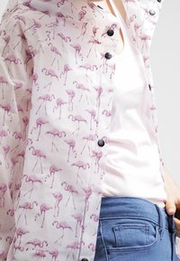 Vit skjorta med knappar och mönster av lila flamingos, bärs över en ljusrosa topp och kombineras med blå jeans.