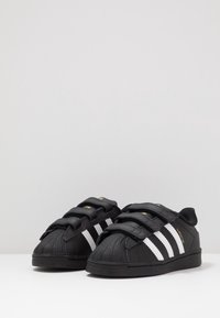 Musta lenkkitossut, joissa on kolme tarranauhaa, kumipohjat, valkoiset raidat ja teksturoitu nahka. Pyöristetty varvas klassisella kuorirakenteella. Kultaiset yksityiskohdat.