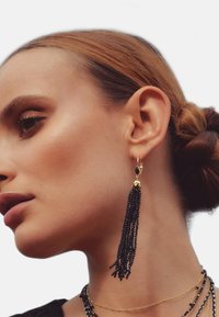 Boucles d'oreilles à franges en perles noires avec quincaillerie dorée. De longues mèches pendent élégamment, ornées d'un sommet décoratif doré. Design élégant et détaillé.