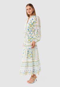 Floral maxi dress in white with blue and green patterns, featuring a cinched waist, long sleeves, and gathered hem; paired with strappy heels.