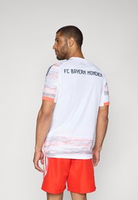 Maglietta bianca del FC Bayern Monaco con motivo grafico rosa e grigio nella parte inferiore, maniche corte e logo sulla schiena. Pantaloni corti rossi completano il look.